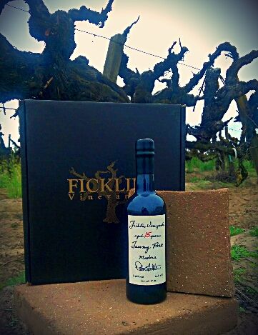 A bottle of Ficklin Vineyards Port stands in front of Ficklin Vineyards logo club members' box with the bare old vines from the vineyard behind them.