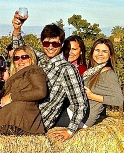 Four guests, with one of them holding a raised glass of Ficklin Port, sitting on straw bales in front of the vineyard.